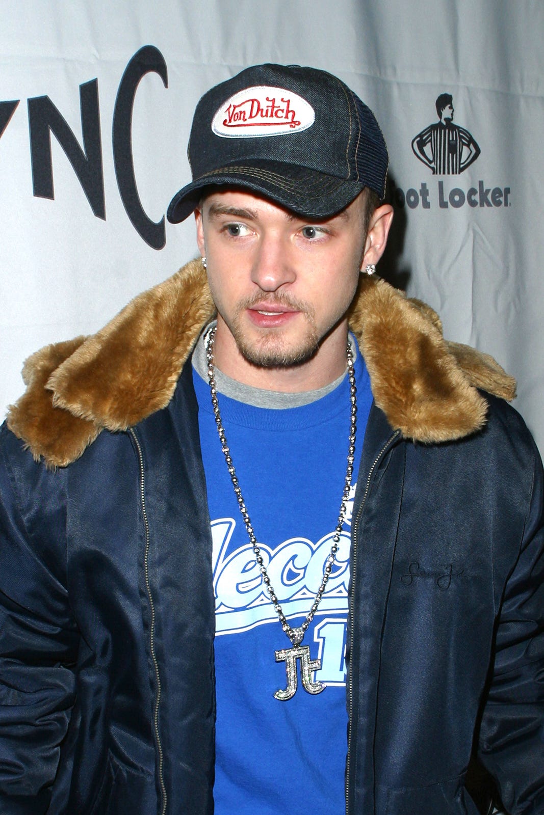 A man wearing a dark Van Dutch cap, a blue shirt, a fur-collared jacket, and a silver chain, stands in front of a step-and-repeat banner with the SYNC and Foot Locker logos.