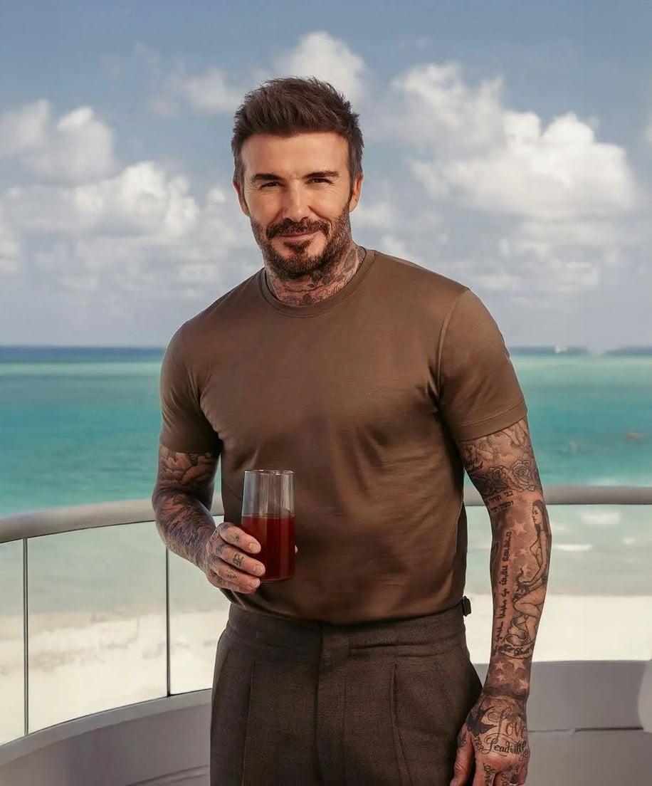 A man with tattoos, wearing a brown t-shirt and gray pants, stands on a balcony by the sea, holding a glass of a dark beverage. The sky is partly cloudy, and the ocean is turquoise in the background.