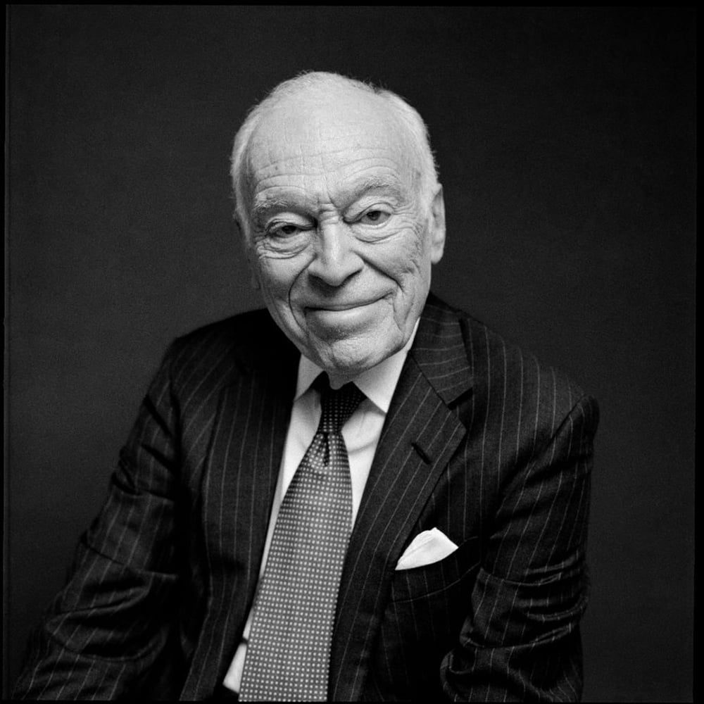 An elderly man in a pinstripe suit, dress shirt, and tie sits against a dark background, smiling warmly at the camera.