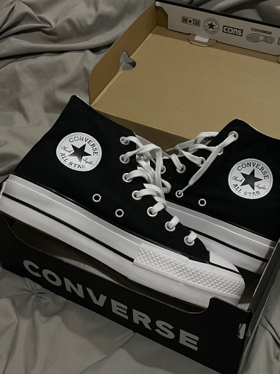 A pair of black and white Converse Chuck Taylor All Star high-top sneakers with white laces, displayed in an open Converse shoe box on a gray fabric surface.