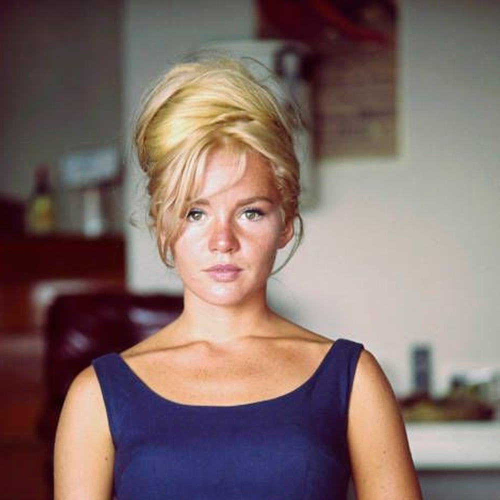 A woman with blonde hair styled in a bouffant updo wears a sleeveless navy blue dress and looks seriously at the camera. The background is an indoor setting with blurred furniture and wall decor.