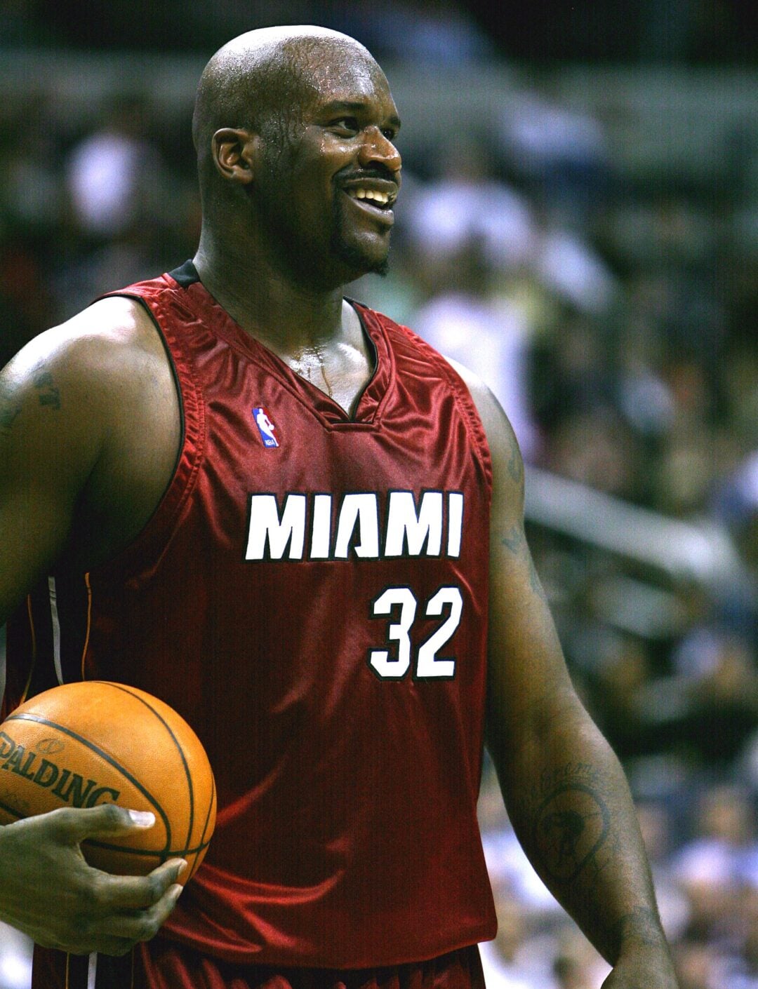 A basketball player wearing a red Miami Heat jersey with the number 32 holds a basketball and smiles during a game. The background shows a blurred crowd in the stands.