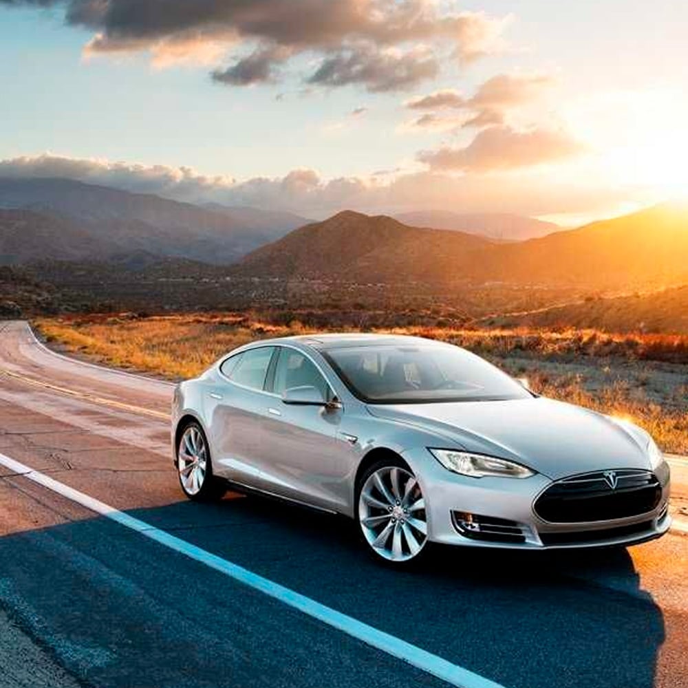 A silver Tesla Model S is parked on an empty road at sunset, with mountains and a partly cloudy sky in the background.