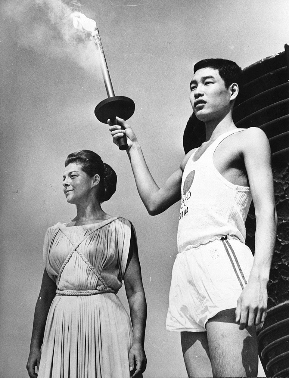 A young man in athletic wear holds a lit Olympic torch beside a woman in a classical Greek-style dress, both standing outdoors against a clear sky.
