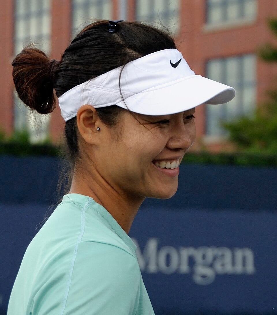A woman with dark hair in a bun, wearing a white Nike visor and a light green shirt, smiles while standing outdoors in front of a blurred building and a blue background with the word "Morgan" visible.