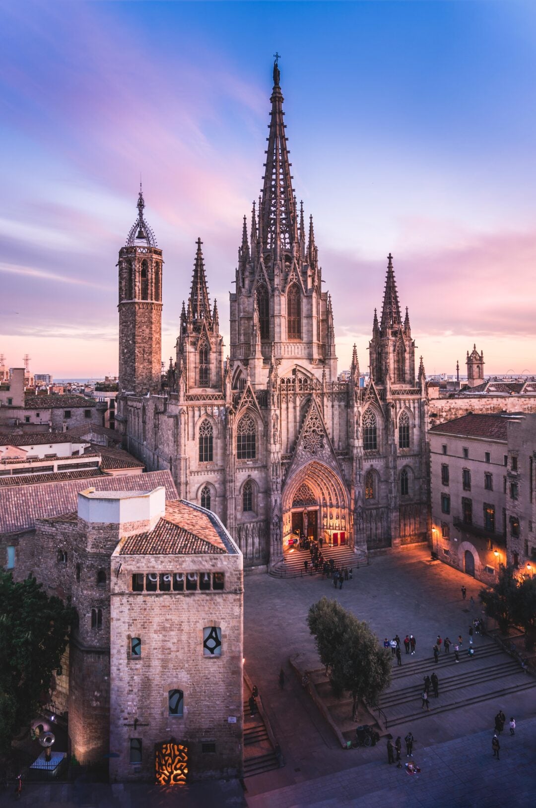 A stunning Gothic cathedral with a tall central spire and ornate details is illuminated at sunset, overlooking a spacious plaza with people and surrounding historic buildings.