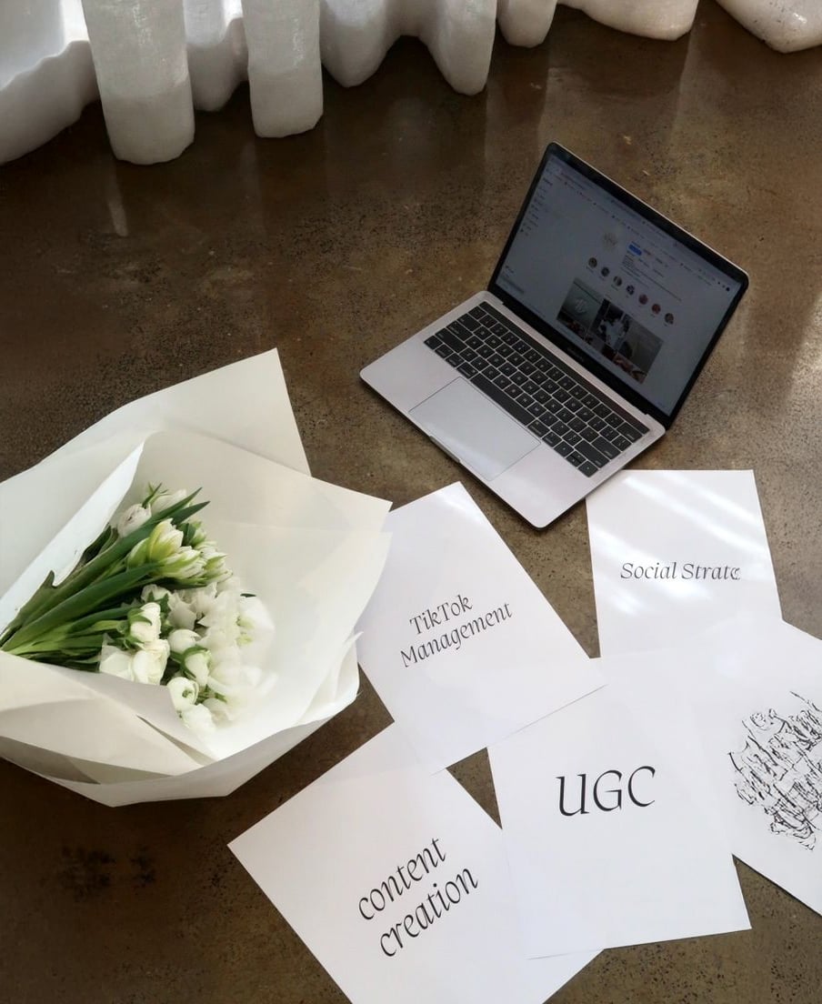 A bouquet of white tulips and a laptop sit on a polished floor beside scattered papers labeled "TikTok Management," "Social Strat," "content creation," and "UGC." Sunlight streams through sheer curtains in the background.