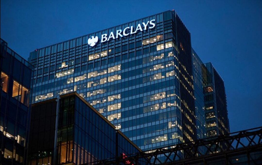 A modern glass office building at dusk illuminated with many interior lights, featuring the Barclays logo at the top. The sky is dark blue and part of a walkway structure is visible in the foreground.