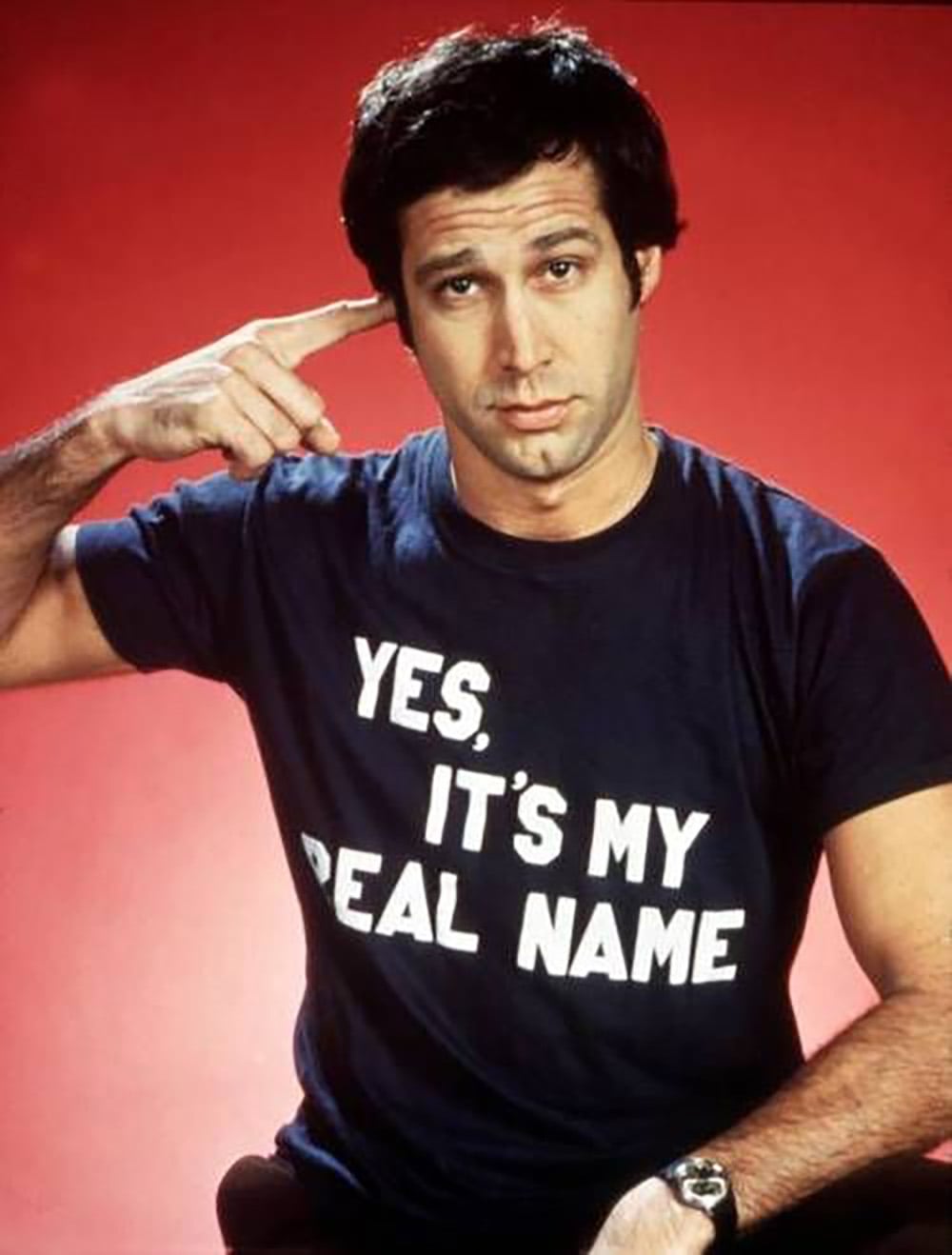 A man with short dark hair points to his head, wearing a navy blue T-shirt that says "YES, IT'S MY REAL NAME" in white letters, standing in front of a red background.