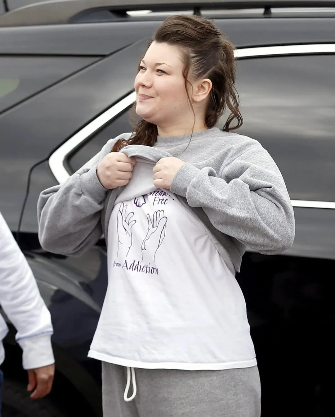 A woman outdoors lifts her gray sweatshirt to show a T-shirt with a design of hands and the words "Free from Addiction." She is smiling and standing near a black car.