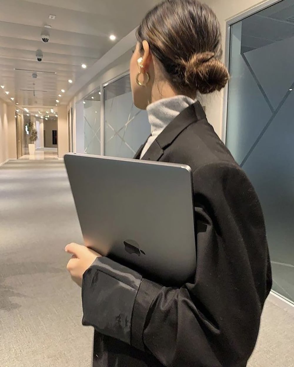 A woman with her hair in a low bun, wearing a black blazer and hoop earrings, holds a closed laptop while walking down a modern hallway with glass doors.
