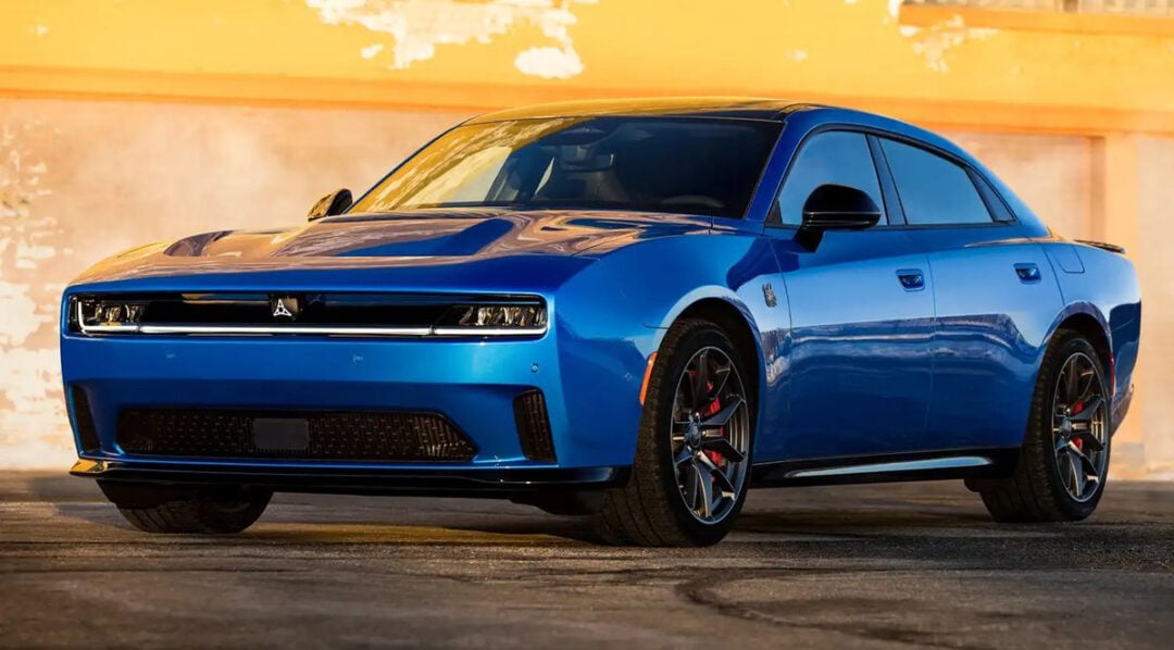 A blue Dodge Charger sports sedan is parked outdoors in front of a weathered yellow and white wall, viewed from the front-left angle under sunlight.