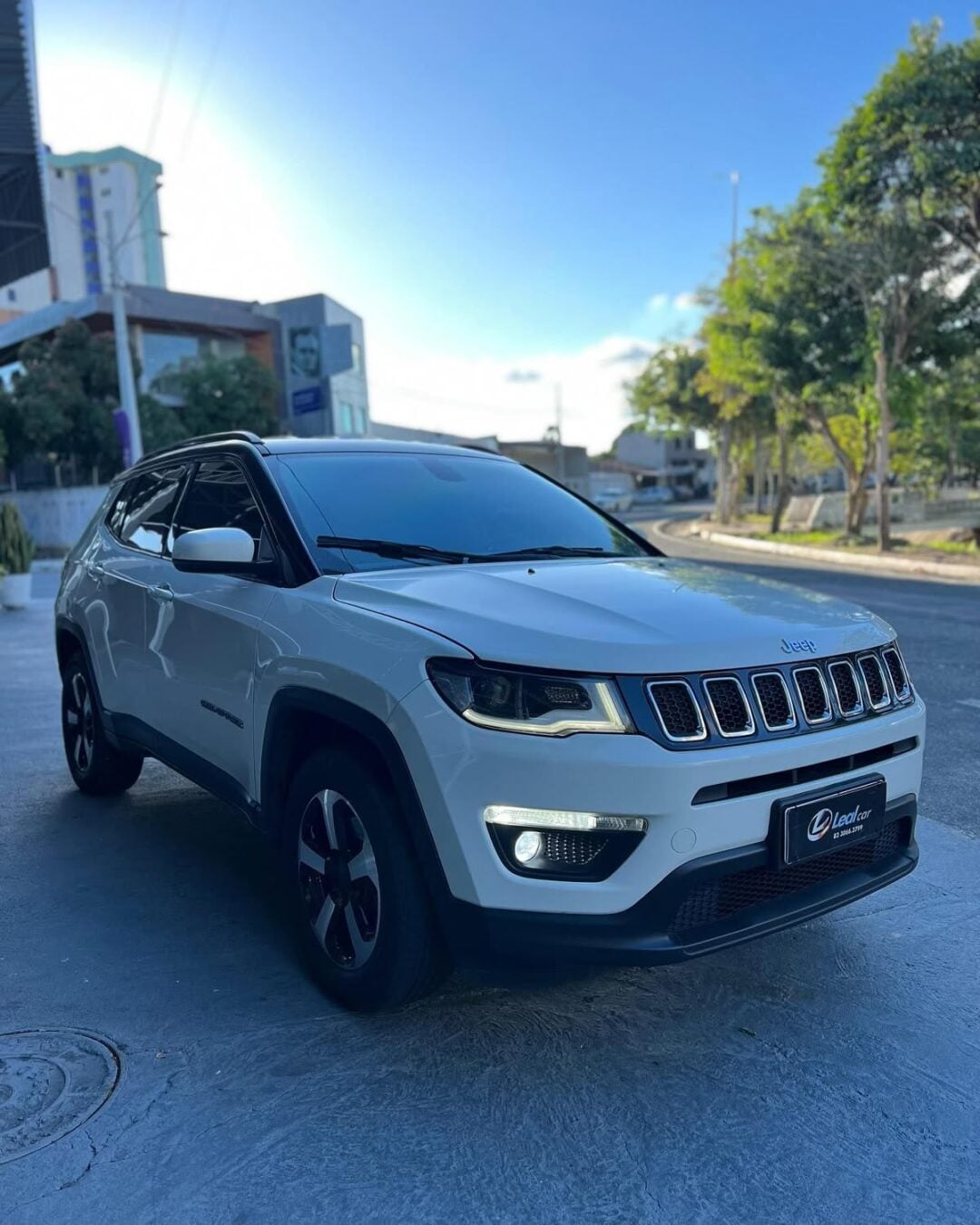 A white Jeep Compass SUV is parked on a city street near a sidewalk, with trees and buildings in the background on a sunny day.