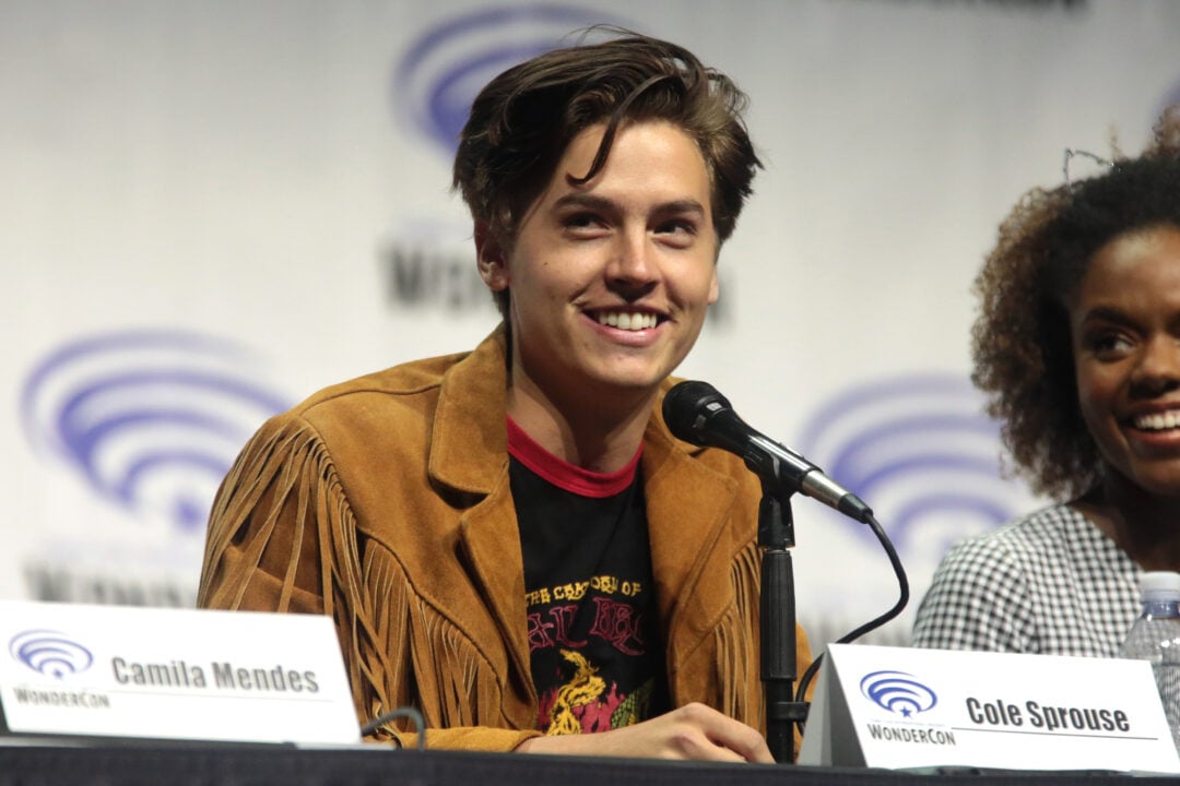 A young man with brown hair wearing a tan fringe jacket smiles while speaking into a microphone at a panel. A woman sits beside him, and name cards on the table read “Camila Mendes” and “Cole Sprouse.”