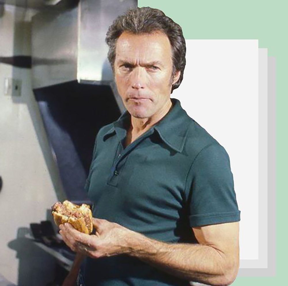A man with short hair in a dark green polo shirt stands indoors, holding a sandwich and looking seriously at the camera. The background is a mix of a kitchen setting and abstract pastel shapes.