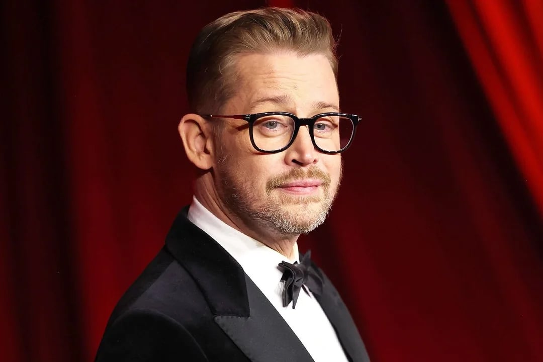 A man with short, light brown hair, glasses, and a trimmed beard wears a black tuxedo with a bow tie, standing in front of a red, softly lit background.