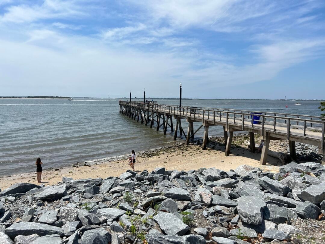 A wooden pier extends over the water from a rocky shore. Two people stand on the sandy beach near the water’s edge under a partly cloudy sky. Distant land and boats are visible across the water.