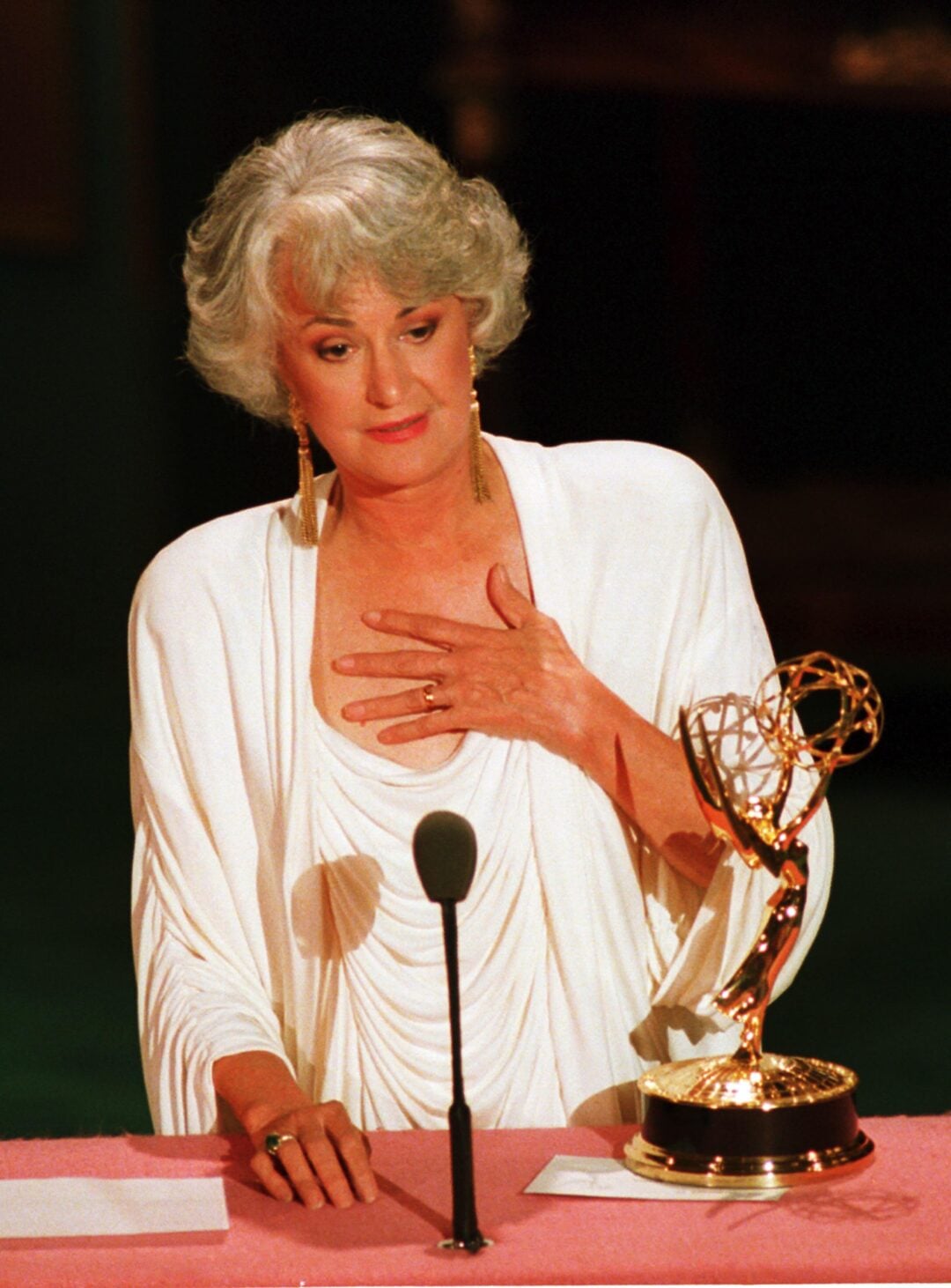 A woman with short gray hair and wearing a white outfit stands at a podium with a hand on her chest, speaking into a microphone, with an Emmy Award trophy in front of her.