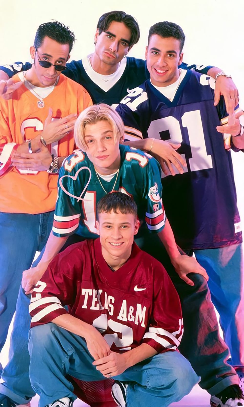 Five young men pose together, all wearing football jerseys in different colors, including orange, green, purple, and maroon. They smile and stand closely against a plain, light background.