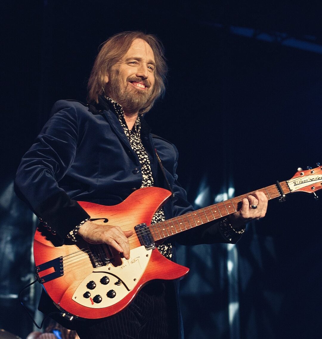 A man with shoulder-length hair and a beard, wearing a dark jacket and patterned shirt, plays a red electric guitar on stage under stage lighting.