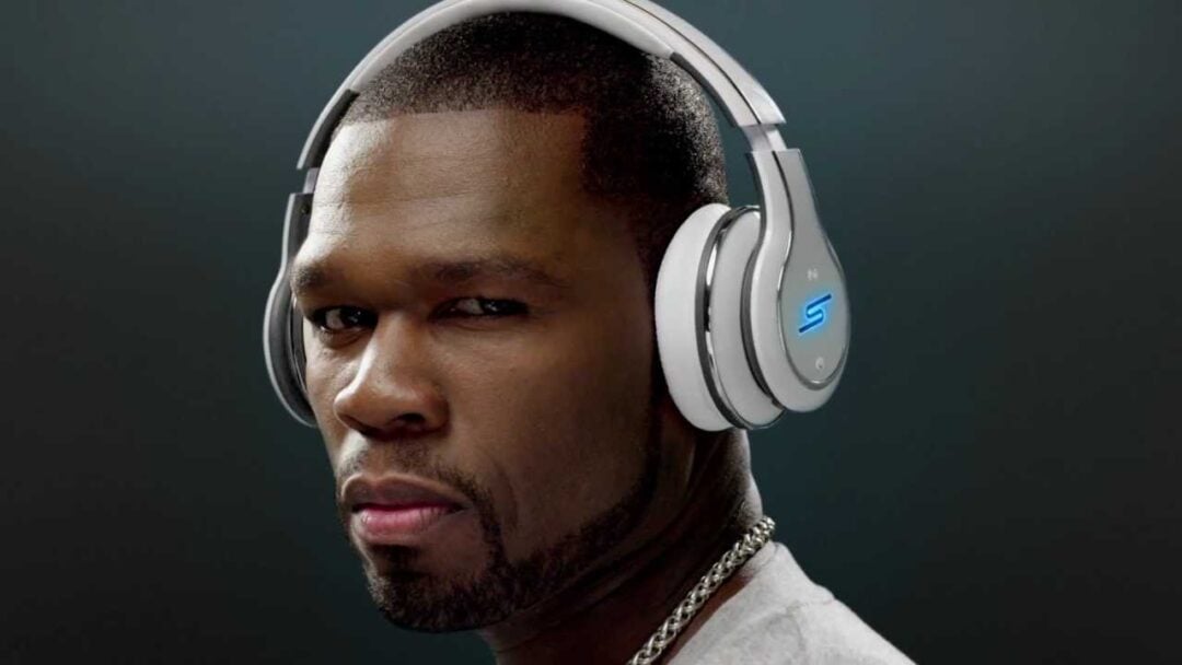 A man wearing large silver over-ear headphones and a silver chain looks directly at the camera with a serious expression. The background is dark and plain.