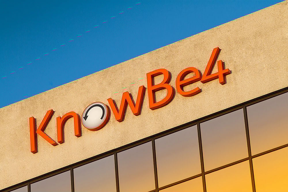 The image shows the KnowBe4 company logo in orange letters on the exterior of a modern building, with a clear blue sky above. The letter "o" has a circular arrow design. The building has reflective windows.