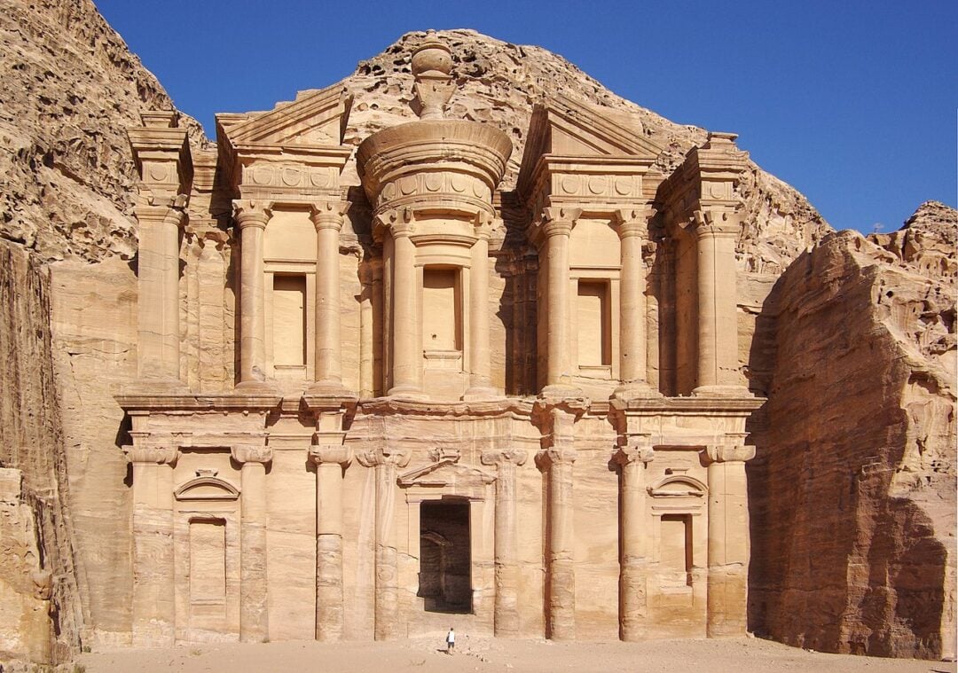 A large ancient stone building carved into a rock face, featuring columns and ornate architectural details, set in a rugged, arid landscape under a clear blue sky.