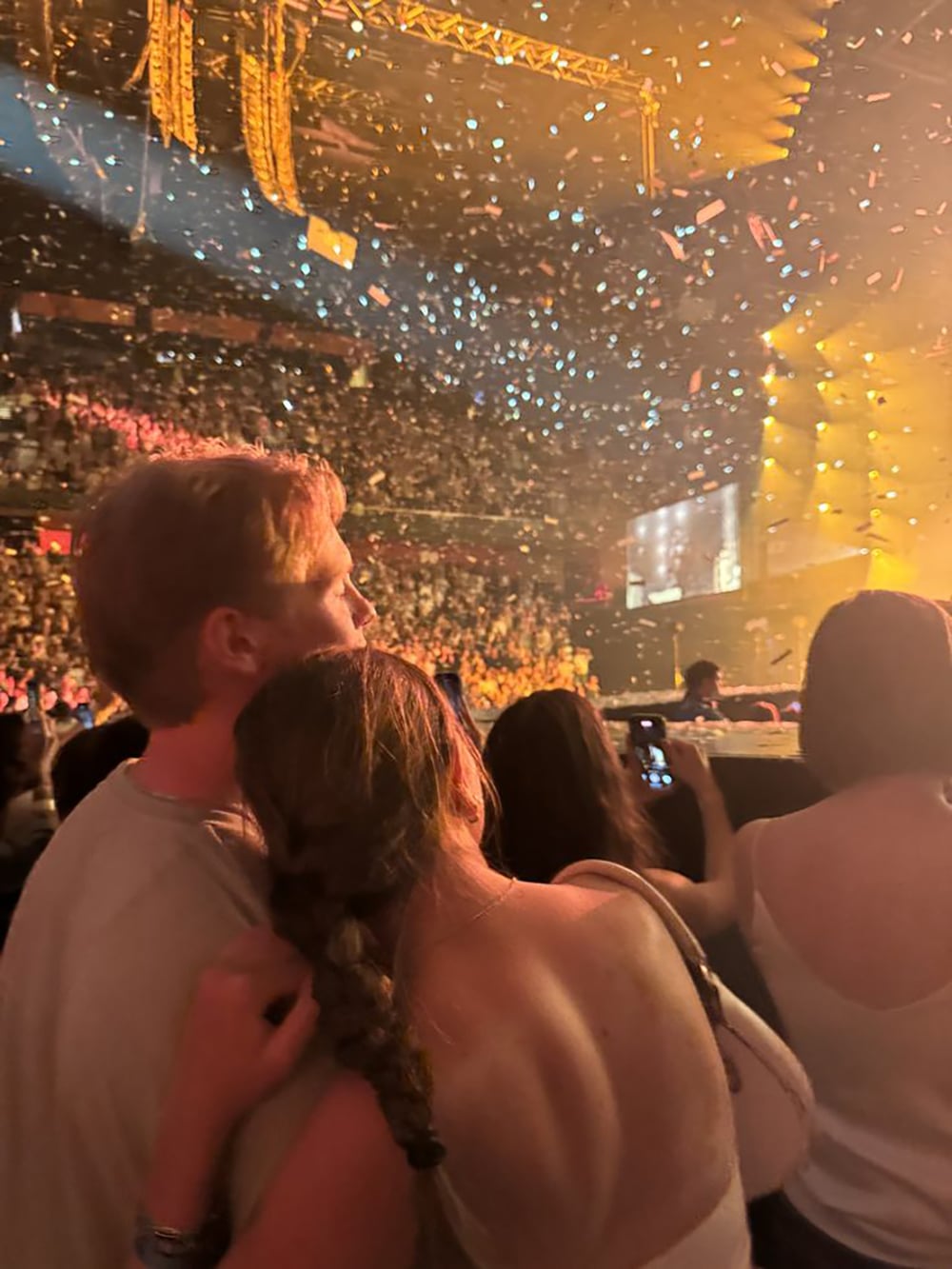 A couple stands close together at a crowded concert, with the woman resting her head on the man's shoulder. Confetti fills the air, bright stage lights shine, and the audience enjoys the lively atmosphere.
