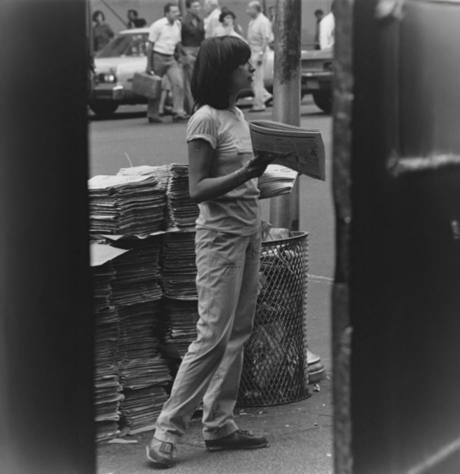 A person stands on a city sidewalk holding a newspaper near a large stack of newspapers by a trash can, with pedestrians and cars visible in the background.