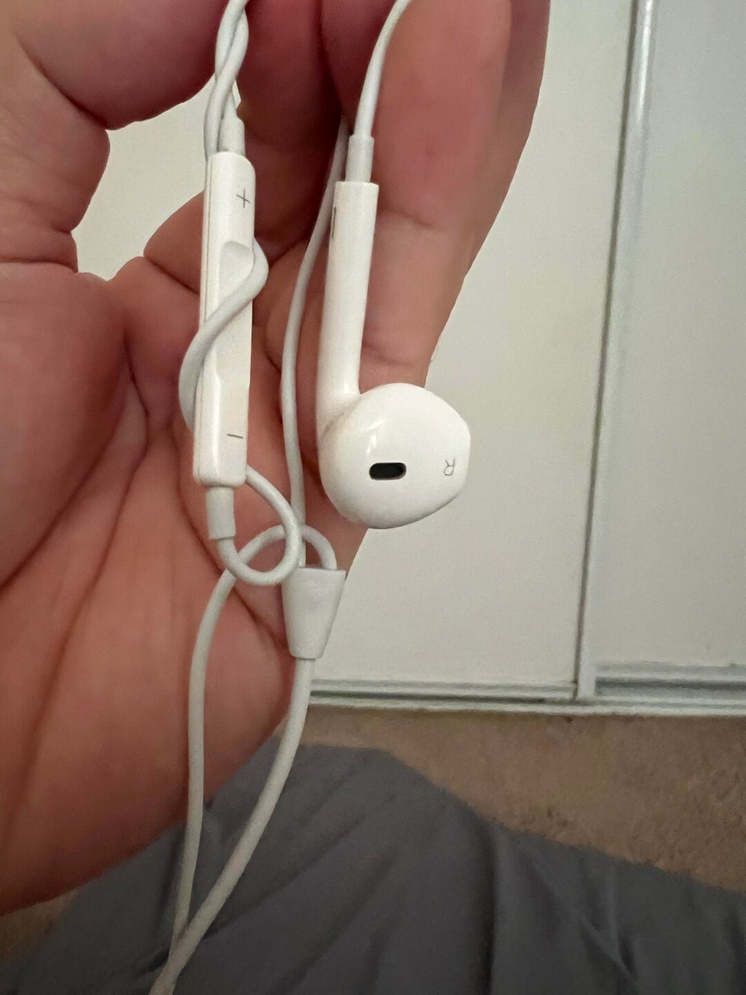 A hand holds a pair of white wired earphones with an inline remote showing plus and minus volume buttons. The background includes a white wall, carpet, and a gray fabric.