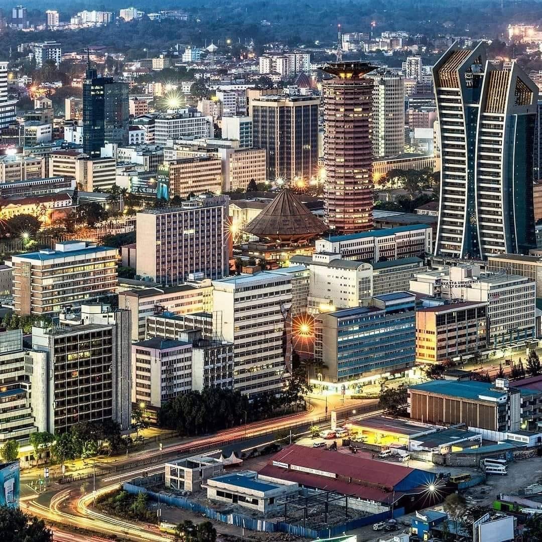 A vibrant cityscape of Nairobi, Kenya at dusk, featuring tall modern skyscrapers, busy streets, and the iconic Kenyatta International Conference Centre with its round tower.