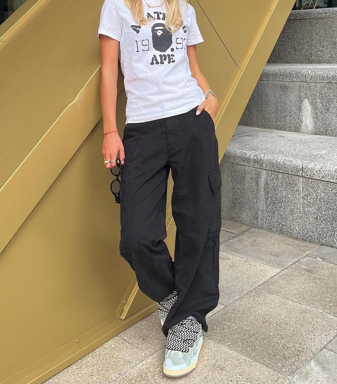 A woman stands outdoors against a metal and stone background, wearing a white graphic T-shirt, black cargo pants, and patterned sneakers. She holds sunglasses and has loose, blonde hair. Her face is not fully visible.