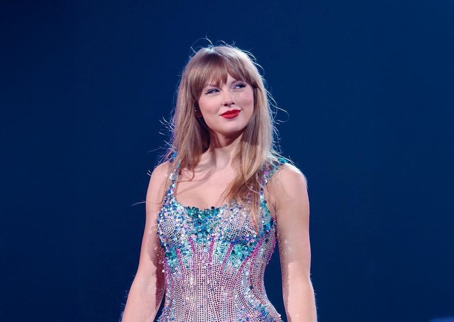 A woman with long blonde hair and bangs stands on stage under blue lighting, wearing a sparkling, colorful dress with sequins and beads. She smiles slightly and looks to the side.