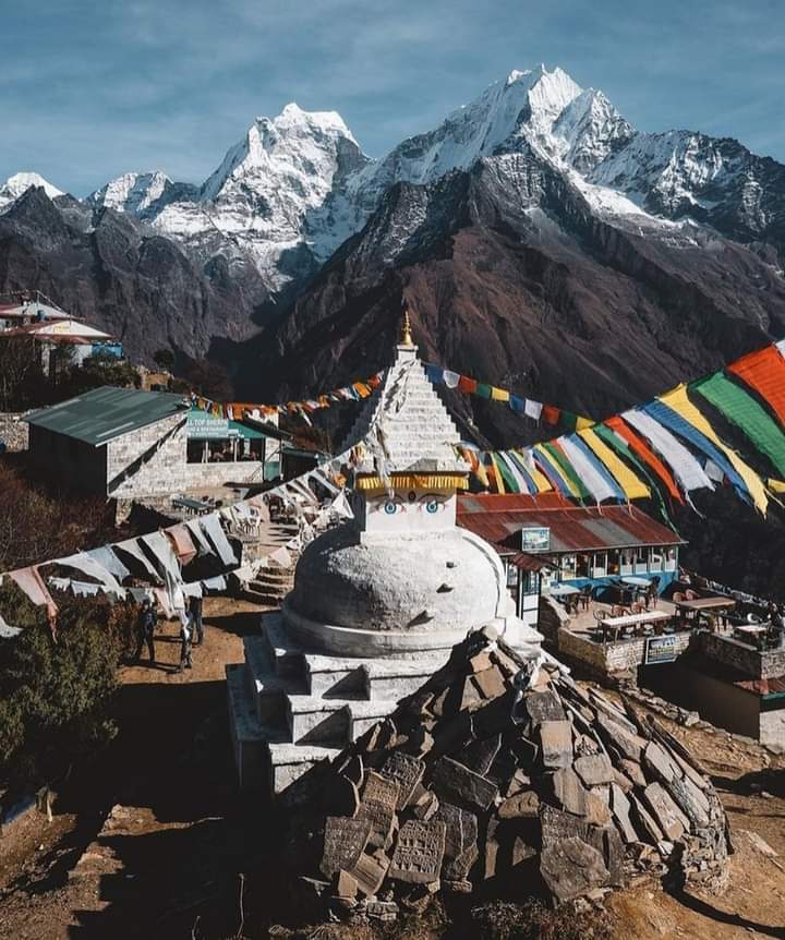 A white Buddhist stupa decorated with colorful prayer flags sits on a mountainside, with snow-capped Himalayan peaks and traditional buildings in the background under a clear blue sky.
