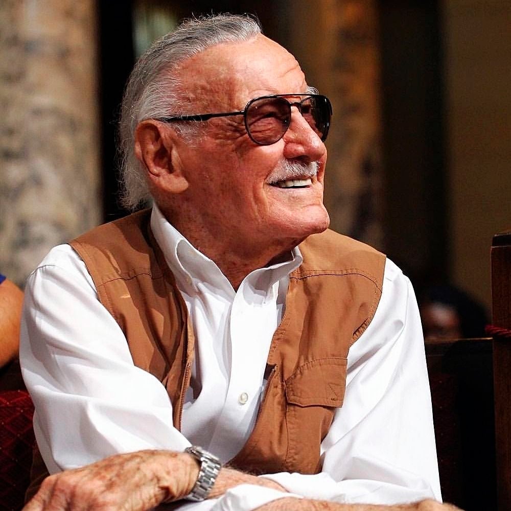 An elderly man with gray hair wearing sunglasses, a white shirt, and a brown vest smiles while sitting indoors.