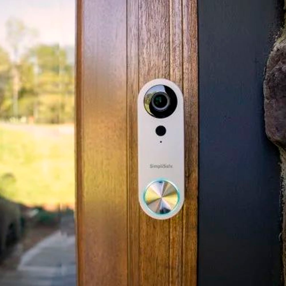 A SimpliSafe video doorbell is mounted on a wooden doorframe, featuring a camera lens, button, and indicator light. The background shows an outdoor scene with greenery.