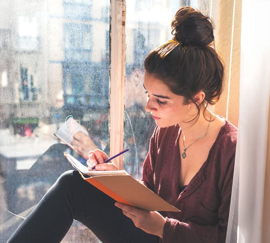 A young woman with dark hair in a bun sits by a sunlit window, writing in a notebook with a pencil. She wears a maroon top and black pants, looking focused and thoughtful.