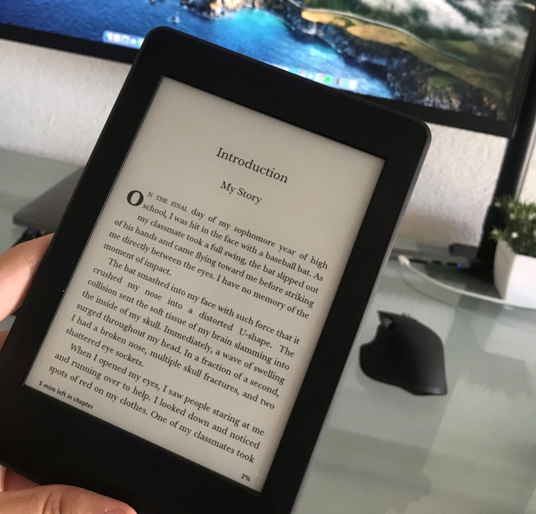 A hand holds an e-reader displaying a book’s introduction page in front of a computer monitor showing a scenic mountain and ocean landscape. The setup is on a glass desk with a mouse, keyboard, and small plant.