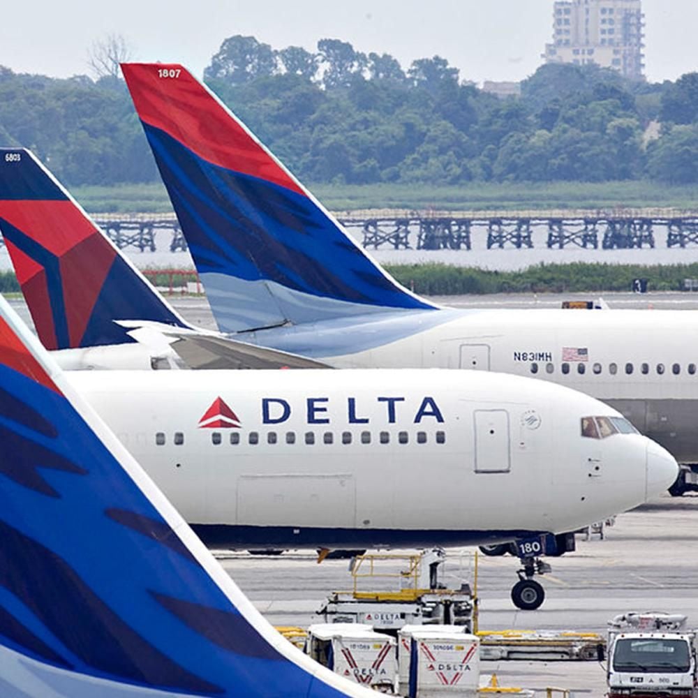 Several Delta Air Lines airplanes are parked at an airport, with their distinctive red, blue, and white tails visible. Ground service vehicles are positioned nearby, and trees and buildings are in the background.