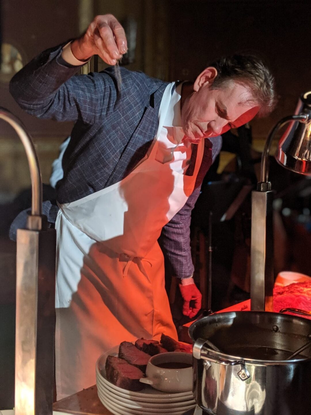 A man in a suit jacket and white apron sprinkles seasoning over sliced meat on a plate, standing under warm lights in a dimly lit setting. A pot and plates are visible on the counter in front of him.