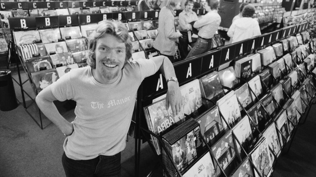 A man with curly hair and a mustache, wearing a "The Manor" t-shirt, smiles while leaning on a rack of vinyl records in a busy record store. Other people browse records in the background.