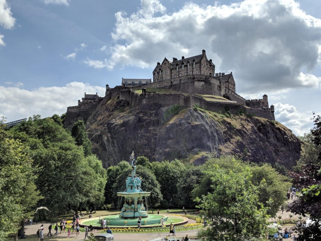 A historic stone castle sits atop a steep, rocky hill, surrounded by lush green trees. Below, a decorative fountain stands in a park with walking paths and people enjoying the sunny day.