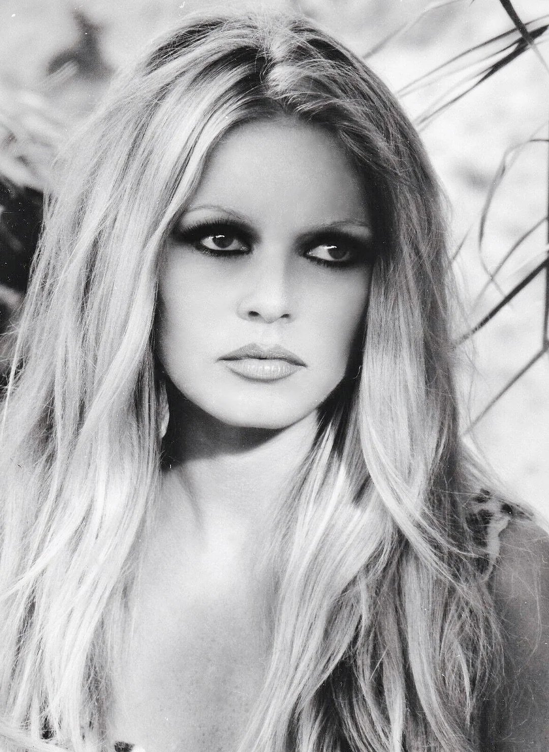 Black-and-white portrait of a woman with long, light hair, dark eye makeup, and neutral expression, looking slightly to the side. Plants and soft background are visible.