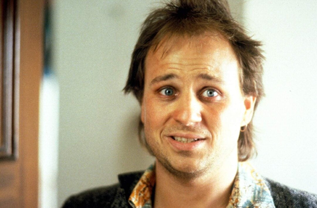 A man with messy brown hair and a scruffy beard stands indoors, looking directly at the camera with a slightly anxious or awkward expression, wearing a patterned shirt and a gray jacket.