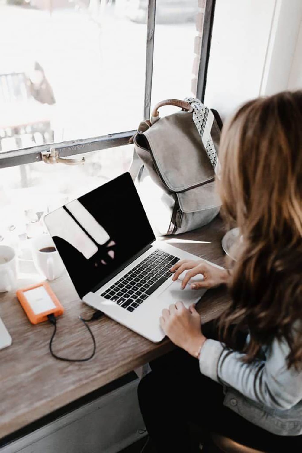 A person with long hair works on a laptop at a wooden desk by a window. A backpack, coffee cup, and external hard drive are on the table. Natural light comes in through the window.