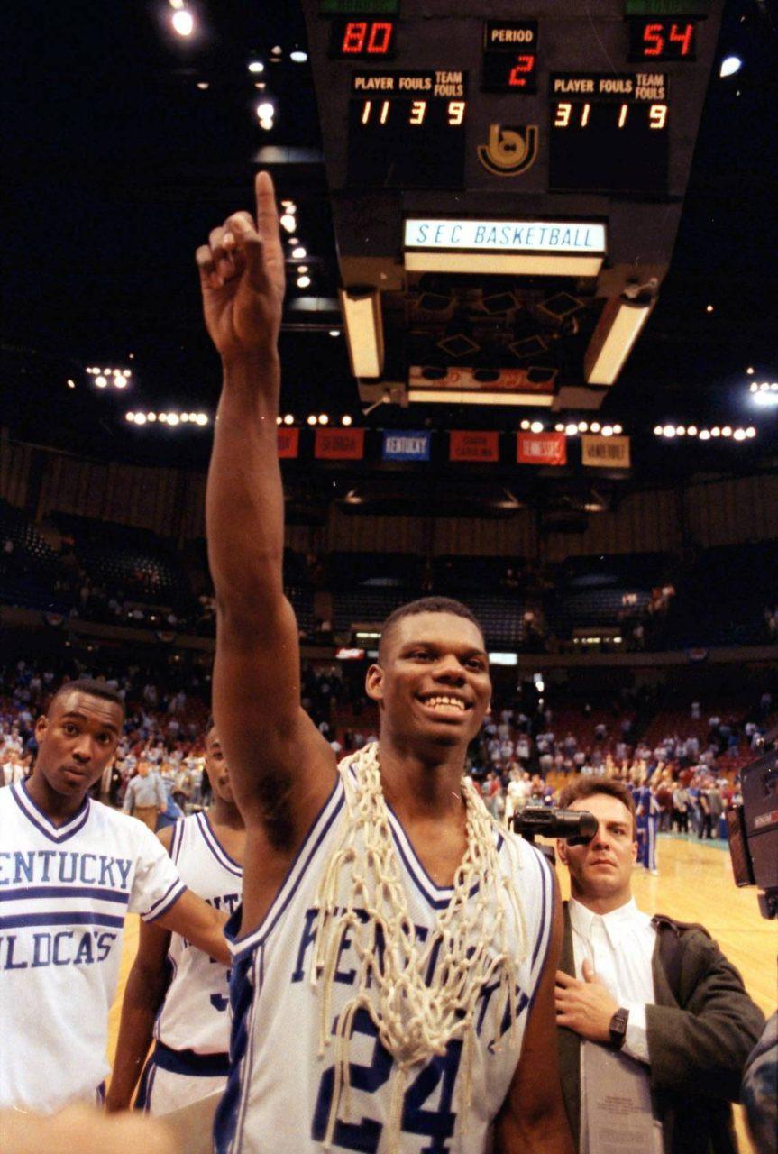 A smiling basketball player wearing a Kentucky Wildcats jersey and a net around his neck raises his finger in victory on a court. The scoreboard above shows a score of 80 to 54. Teammates and people surround him, celebrating.