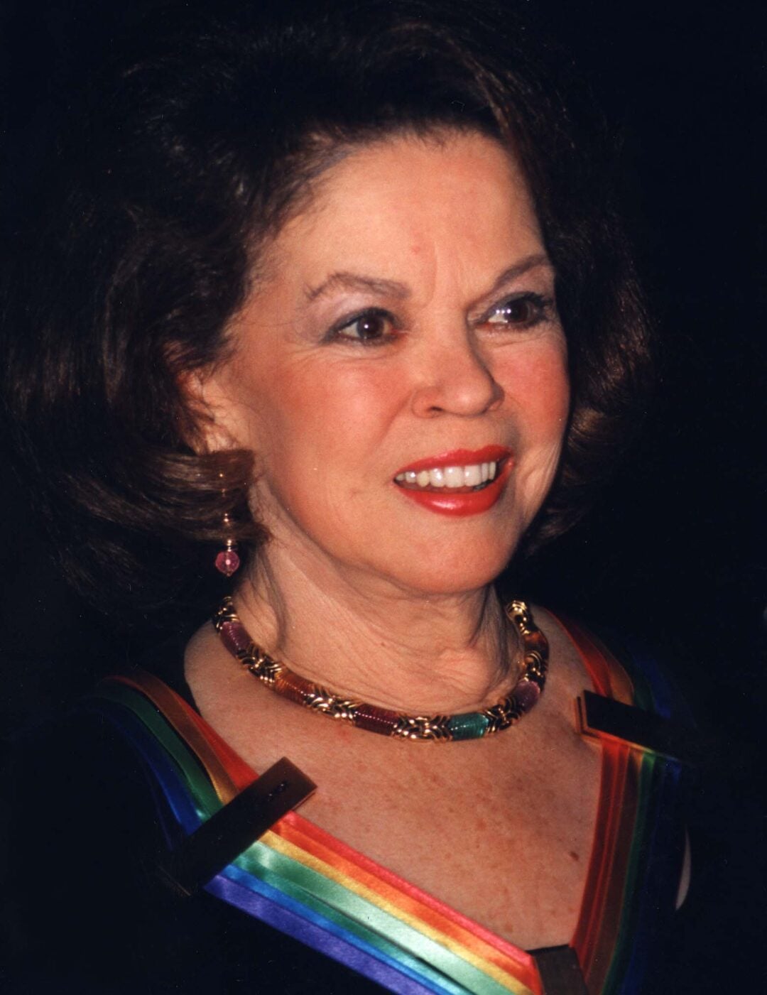 A woman with shoulder-length brown hair smiles, wearing a black dress with a rainbow-colored collar, a gold necklace, and earrings, against a dark background.