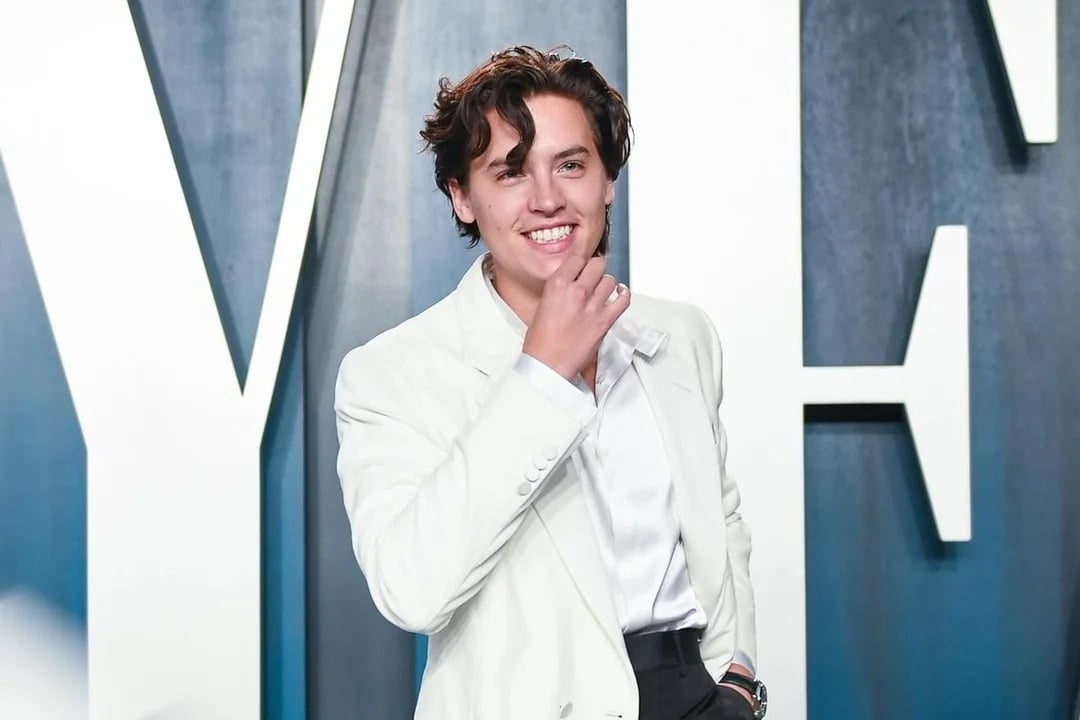 A young man with wavy brown hair, wearing a white suit jacket and satin shirt, smiles with his hand touching his chin while standing in front of a large, stylized "V" and "A" background.