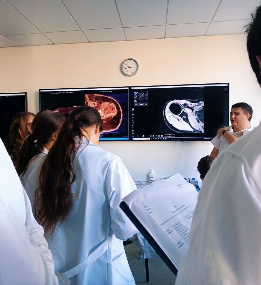 Medical students in white lab coats observe and take notes while an instructor explains medical scans displayed on large screens in a classroom setting. A clock on the wall shows the time as 11:12.