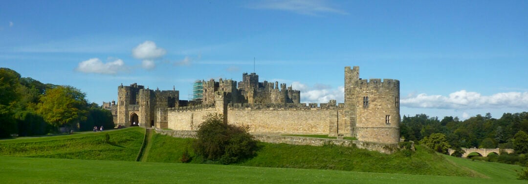 A large stone castle with tall towers and fortified walls stands on a grassy hill under a blue sky with scattered clouds, surrounded by trees and greenery.